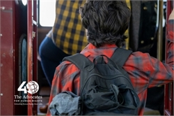 [ai] A person in a red and black plaid shirt with a backpack is standing near a bus entrance, watching as another individual wearing a yellow and black checked shirt steps inside the bus.
